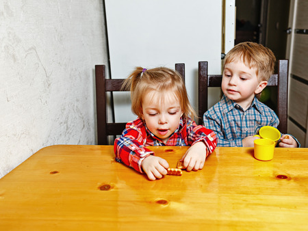 Happy brother and sister sitting at the kitchen table. Little cute girl holding in her hands a cookie, and a boy holding a cup. Love, Family, Friendshipの写真素材