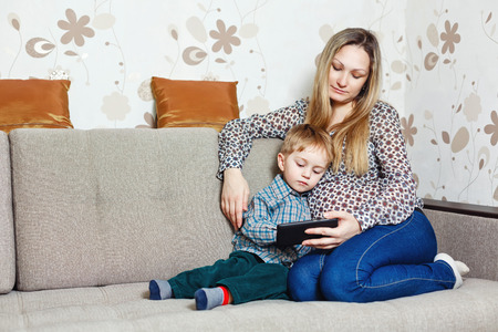 Mother and son playing on tablet, smiling, sitting on the couch. Happy family leisure, modern people.の写真素材