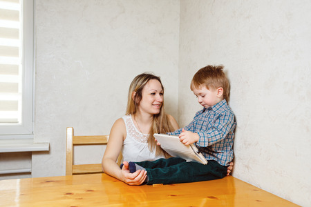Mother and child play on a Tablet PC in the kitchen. Family leisure, modern, young and technology. Boy holding a tablet, and the mother smiles at him.の写真素材