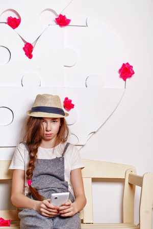 Teenage girl in a hat sitting on a bench. Sad teenager holding a cell phone. The concept of youth culture.の写真素材