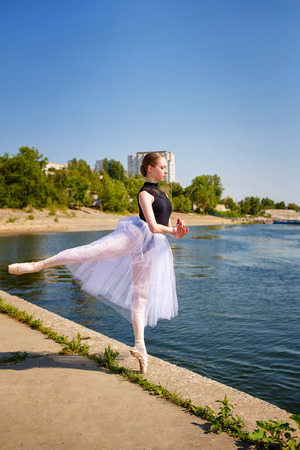 Young slim ballerina in tutu dancing on the riverbank. Summer day. The concept modern ballet. Performance on the street. Arabesqueの写真素材