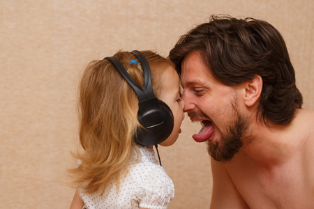 Little cute girl listening to music with big headphones. Daughter and father show each other tongue. Happy family.の写真素材
