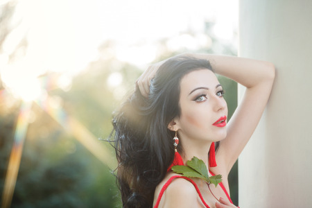 Attractive brunette girl in the red dress. She straightens her hair. Sun glare in her hair. Sunset. Art Deco and Art Nouveau. The concept of retro fashion.の写真素材