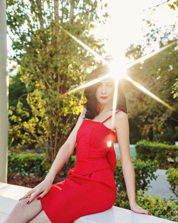 Attractive brunette girl in the red dress. Portrait at sunset. Sun glare in her hair. Art Deco and Art Nouveau. The concept of retro fashion.の写真素材