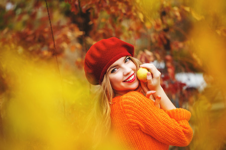 Pretty girl in a beret and a sweater in autumn park, holding a ripe apple and smiling. Girl enjoying fresh air. The girl white smile. The concept of modern youth fashion.の写真素材
