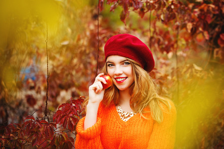 Nice girl in a beret and a sweater in autumn park, holding a ripe apple and smiling. The girl white smile. The concept of healthy teeth.の写真素材
