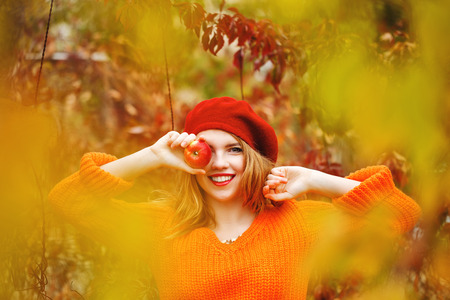 Pretty girl in a beret and a sweater in autumn park, holding a ripe apple and smiling. Girl hiding eye for fruit. The girl white smile. The concept of healthy teeth.の写真素材