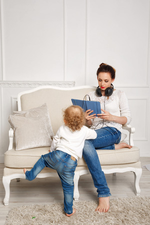 Mother and daughter at home. Baby daughter climbing onto the couch. Mother listens to music on headphones and chtiaet news on tablet pc. Girls dressed in white sweaters. Family timeの写真素材