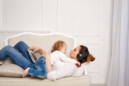 Mother and daughter at home. Little daughter hugging her mother. Mother listens to music in headphones and holding a tablet pc. Girls dressed in white sweaters. Family timeの写真素材
