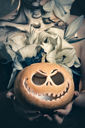 Jack-o'-lantern. Pumpkin close up in the hands of women. Young attractive girl with creative make-up for Halloween. Mysterious and frightening image of lilies. Witchcraft. Horrible.の写真素材