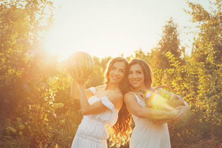 Two cute sisters in long white dresses hold in hands of the crop. Girl holding a basket of ripe fruits and vegetables, and the other girl is holding a watermelon. A bountiful harvest. Agriculture. The setting sun. Soft focusの写真素材
