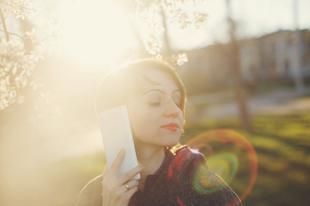 Cute girl with the phone close-up portrait on a background of cherry blossoms and the setting sun. The solar lens flare.の写真素材