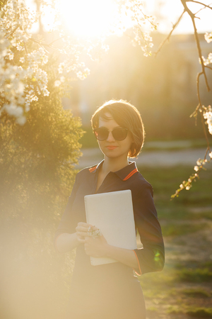 Cute girl holding a laptop in hand. Portrait against the setting sun and cherry blossoms. She is wearing sunglassesの写真素材