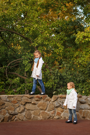 Cute little sisters in the autumn park. The older girl is on the stone fence. Family time. Sport games.の写真素材