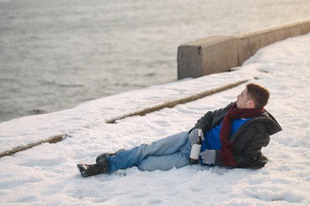 Man walking with a bottle of wine. He is dressed in winter coat, scarf and gloves. Man was drunk. He lies on the snow on the quay. Winter depression.の写真素材