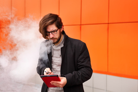 Young hipster man with a beard and glasses smokes an electronic cigarette. He holds a tablet pc. Youth fashion. Delicious pairs. Man makes startup. Internet business.の写真素材