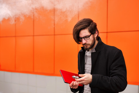 Young hipster man with a beard and glasses smokes an electronic cigarette. He holds a tablet pc. Youth fashion. Delicious pairs. Without nicotine liquid.の写真素材
