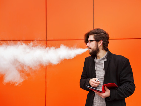 Young hipster man with a beard and glasses smokes an electronic cigarette. He holds a tablet pc....