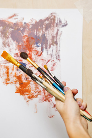 Hand of an artist with brushes on a background of an abstract painting. Synthetic and natural brushes. Art supplies. Creative expression tools.の写真素材
