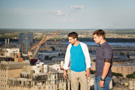 Two roofers on the roof of a high-rise building admire the view of the city. Courage and adrenaline. The men are looking down.の写真素材