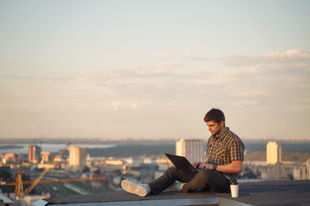 Hipster man with a laptop is drinking coffee on the roof. Freelancer at work.の写真素材