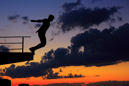 Silhouette of a man against a background of clouds and golden sunset. He jumps from the edge of the roof.の写真素材