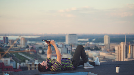 A hipster guy makes a selfie on the roof of a high-rise building. The video blogger broadcasts from a new height.の写真素材