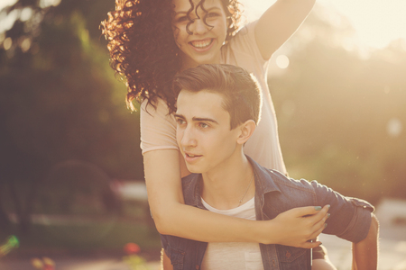 Teenagers are walking in the city park. He rolls her on the shoulders of piggyback. Couple in love. Romantic first date. Carefree youth.の写真素材