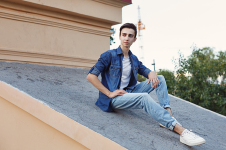 A young guy is sitting on the roof. He is wearing a jacket and jeans. Hipster city fashion.の写真素材