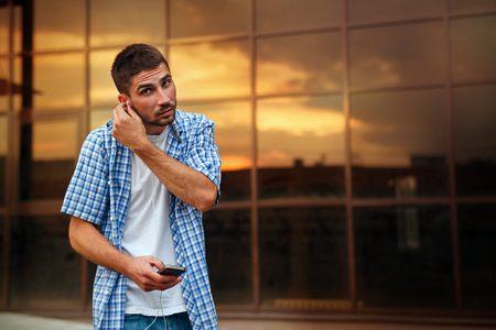 A hipster man listens to music on headphones. He is dressed in a plaid shirt and a white T-shirt, holding a cell phone.の写真素材