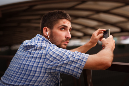 A hipster man listens to music on headphones. He is dressed in a plaid shirt and a white T-shirt, holding a cell phone. The guy is waiting for the train on the feather.の写真素材