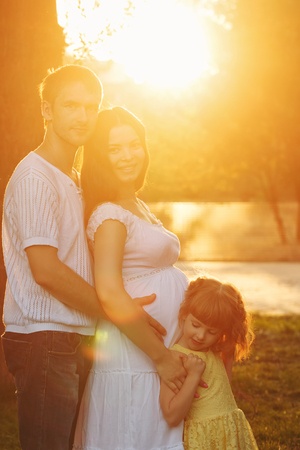 Family. Pregnant mother, father and daughter. Father hugs mother, and mother hugs daughter. Family portrait at sunset. Happiness of parenthoodの写真素材