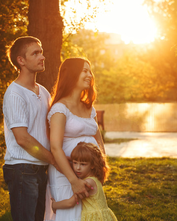 Family. Pregnant mother, father and daughter. Father hugs mother, and mother and daughter hug each other. Family portrait at sunset. Happiness of parenthoodの写真素材