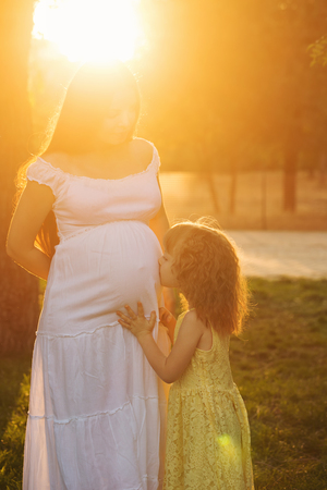 Family. A pregnant mother hugs her little daughter. The daughter kisses her belly. Family portrait at sunset. Happiness of parenthoodの写真素材