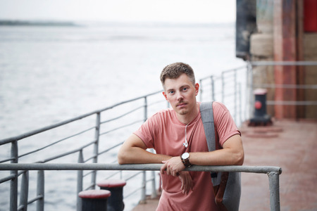 The guy on the pier is waiting for the ship. Ferry crossing. He is wearing a T-shirt. He has a backpack and headphones with him. Youth fashion.の写真素材
