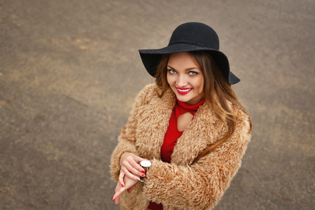 Young attractive girl outdoors in winter. She is wearing wide-brimmed hat and fur coat. The girl is standing next to fir-tree. She looks at clock how much time nowの写真素材
