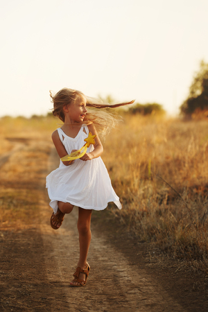 A little girl in a white dress and sandals runs along a country road. She holds a toy in her hands. Happy childhood. Summer vacationの写真素材