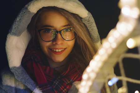 Young attractive hipster girl wearing glasses is walking by the mall on holiday. She smiles. Portrait at night next to the garlands.の写真素材