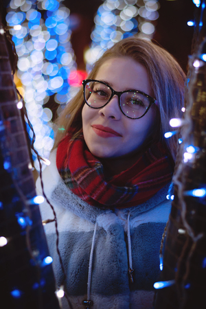 A young attractive girl in glasses and a warm scarf walks at the city fair. She smiles. Portrait at night among garlands.の写真素材