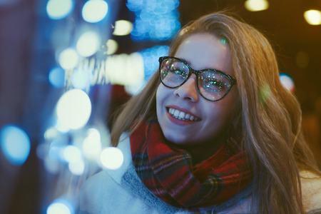 A young attractive girl in glasses and a warm scarf walks at the city fair. She is smiling. Portrait at night among garlands.の写真素材