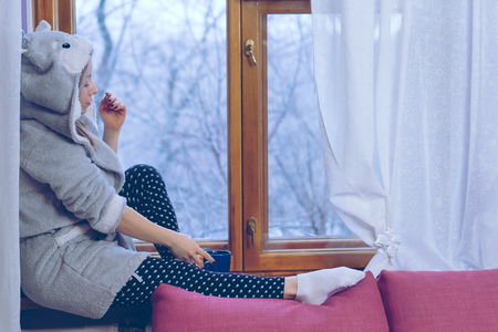 A nice girl is drinking tea sitting on the windowsill at home. She holds a mug with a warming drink in her hands. It's freezing outside.の写真素材