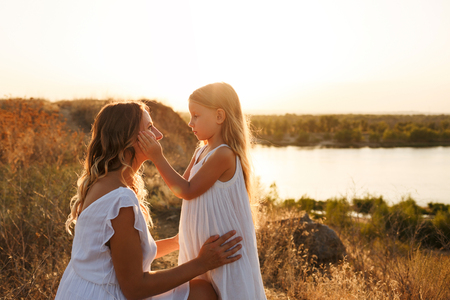 Family, mother and daughter are standing on the river bank. A little girl and mother look into each other's eyes. Girls in white dresses. They are blondes. Family time together.の写真素材