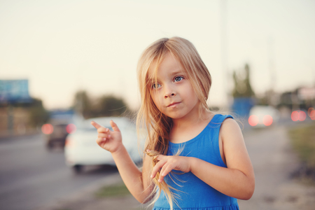 A little girl in a blue dress is standing by the busy highway. In the background, the machines are in a defocus.の写真素材