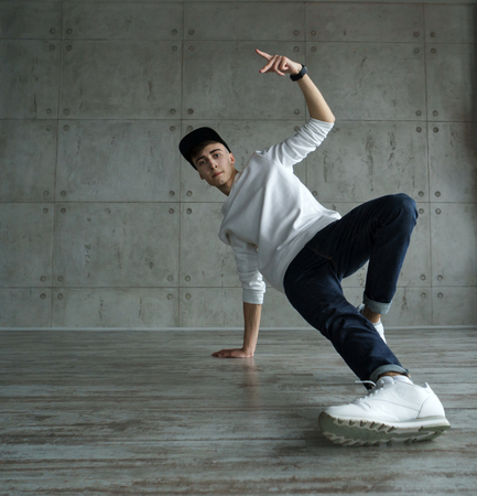 Teenage boy in white sweater and baseball cap, jeans and sneakers makes breakdance in the dance studio. Dynamics of modern dance movementの写真素材