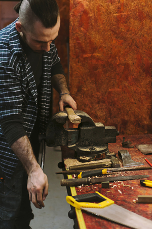 Man works in carpentry workshop. He fixes wooden handle in vice. Different tools are on workbench....