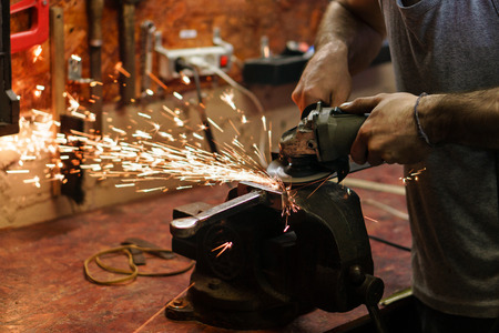 Blacksmith polishes crossguard of sword. Man is working in workshop. He holds a grinder in his...