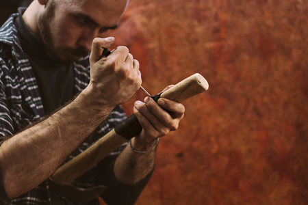 Man working in carpentry workshop. He pierces leather winding on wooden handle of ax. Men at work. Hand work.の写真素材