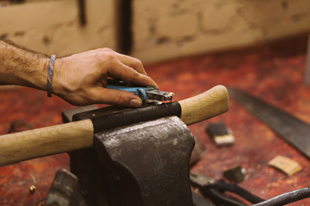Man working in carpentry workshop. He fastens leather winding on wooden handle of ax. Men at work. Hand work.の写真素材