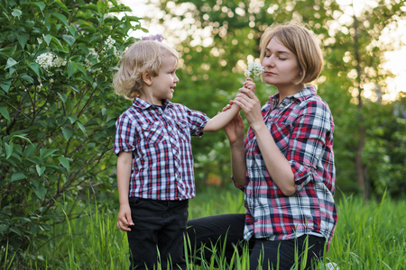 Mother and son in plaid shirts. Woman and boy are standing by flowering bush of lilac. She smells flowers. Family time together.の写真素材
