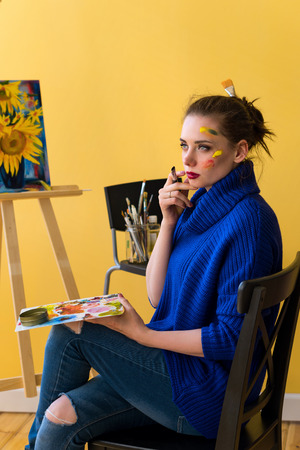 Girl artist paints sunflowers oil paints on canvas. She is wearing blue sweater. Woman is holding brush and palette with paints. She dreams.の写真素材